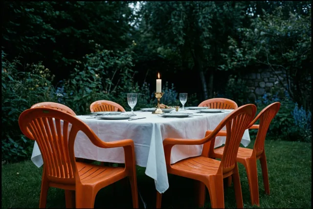Een gedekte tuintafel met een wit tafelkleed, oranje plastic stoelen en een brandende communiekaars in een gouden houder staat tussen donkergroen gebladerte in het avondlicht.