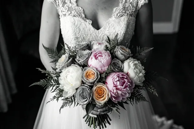 Een bruid in een kanten trouwjurk houdt een boeket pioenrozen en rozen vast, met roze en perzikkleurige bloemen in kleur tegen een zwart-witte achtergrond.