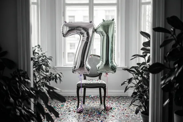 Folieballonnen in de vorm van 7 en 1 hangen aan een stoel, met gekleurde confetti op de vloer in een verder grotendeels zwart-witte kamer.
