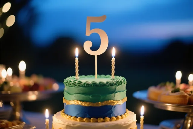 Een taart met meerdere verdiepingen, groen, blauw en wit glazuur, gouden accenten, een groot cijfer 5 en brandende kaarsjes staat op een buffet onder een schemerlucht met onscherpe lichtjes.