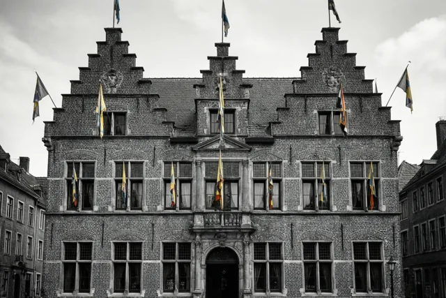Een zwart-witbeeld toont een bakstenen stadhuis met trapgevels en veel ramen onder een bewolkte hemel, waarbij alleen de carnavalsvlaggen gekleurd zijn.