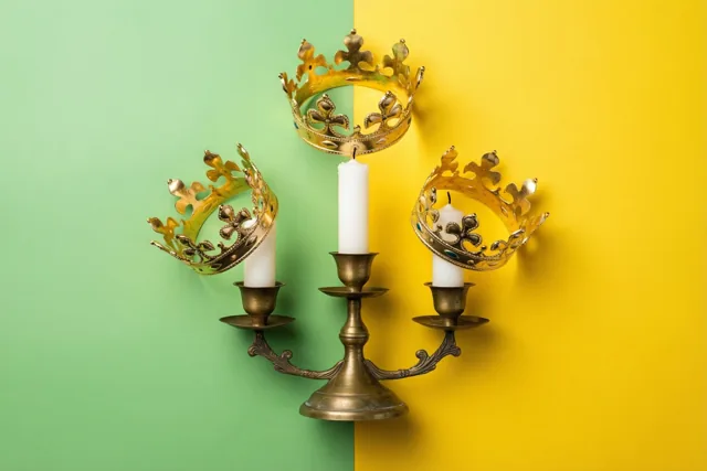 Een messing kandelaar met drie witte kaarsen, elk met een dun gouden kroontje bovenaan, staat tegen een achtergrond met een groen en een geel vlak.
