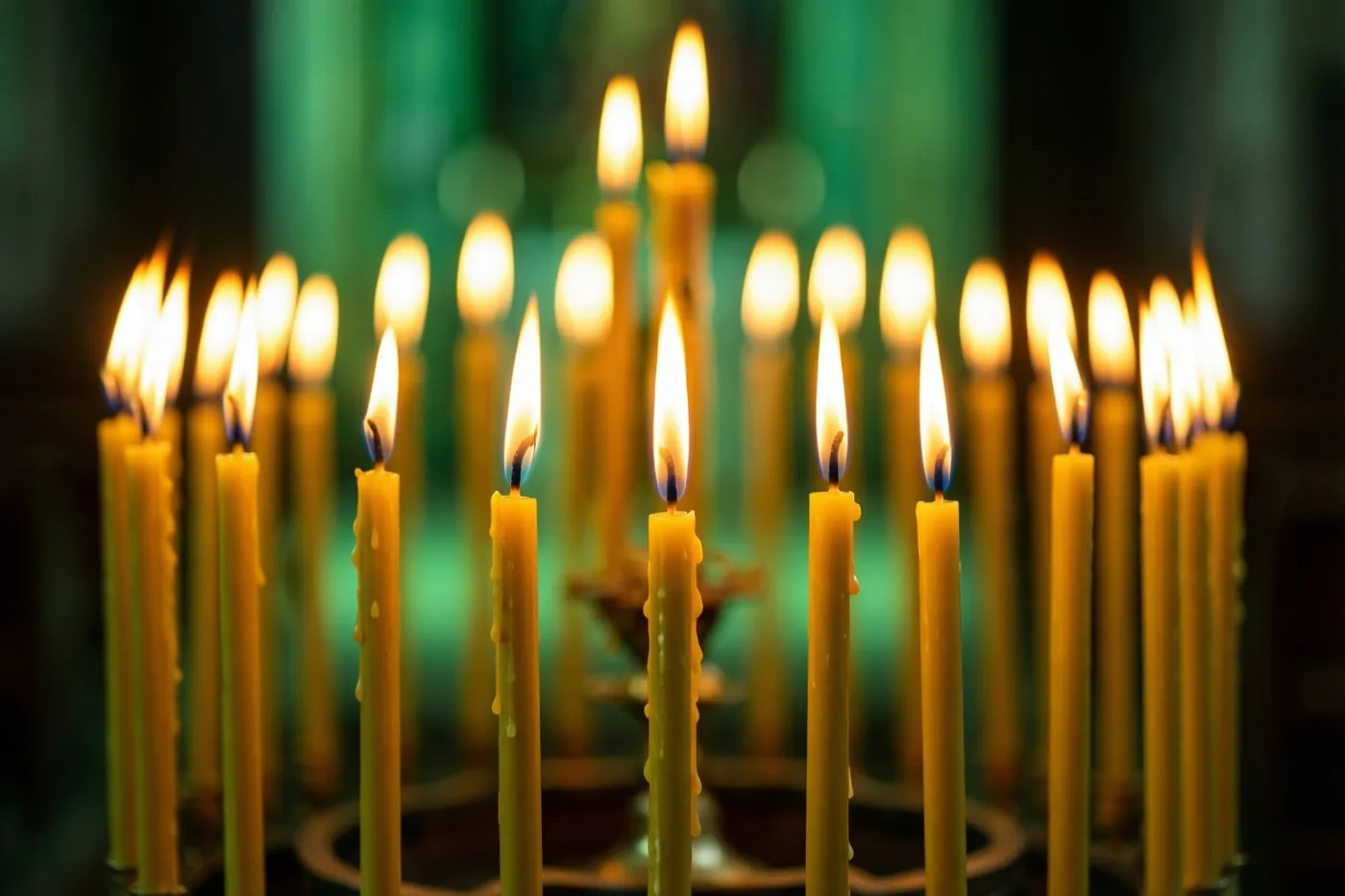 Een rij brandende gele kaarsen met smeltend kaarsvet staat in een donkere kerk tegen een onscherpe groene achtergrond.