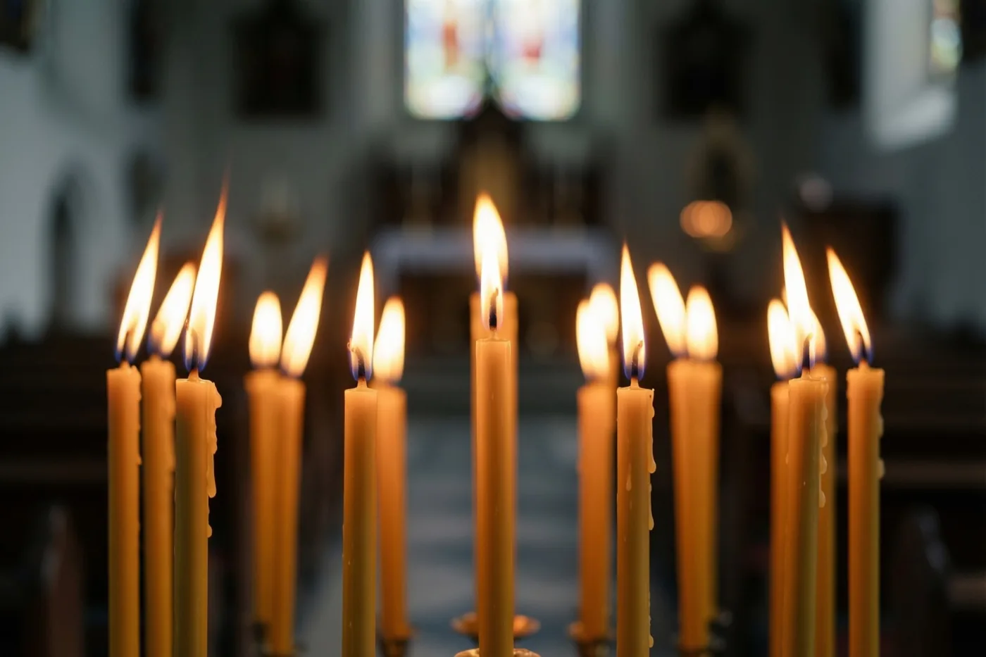 Een rij brandende gele kaarsen met onscherpe vlammen staat in een donkere kerk, met wazige glasramen en banken op de achtergrond.