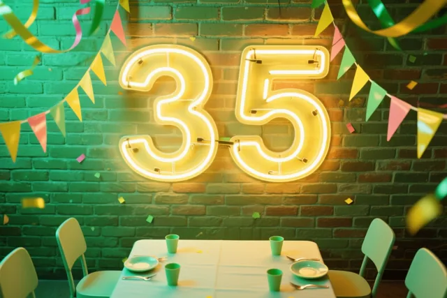 Een warmgeel neongetal 35 hangt aan een bakstenen muur boven een feesttafel met kleurrijke serpentines en confetti.