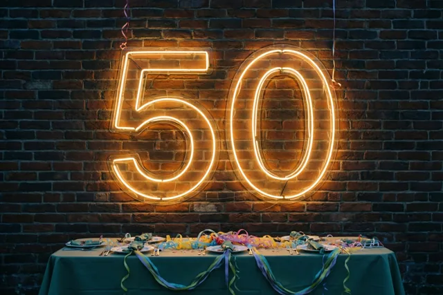 Een groot geel neongetal 50 hangt aan een donkere bakstenen muur boven een tafel met een groen tafelkleed, kleurrijke serpentines en borden.