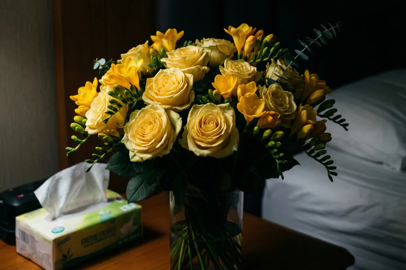 Gele rozen en fresia’s staan in een heldere glazen vaas op een houten nachtkastje naast een doos zakdoekjes in zacht warm licht.