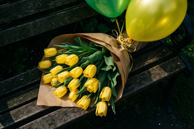 Gele tulpen in bruin papier liggen met groene en gele ballonnen op een donkerhouten tuinbank in fel zonlicht.