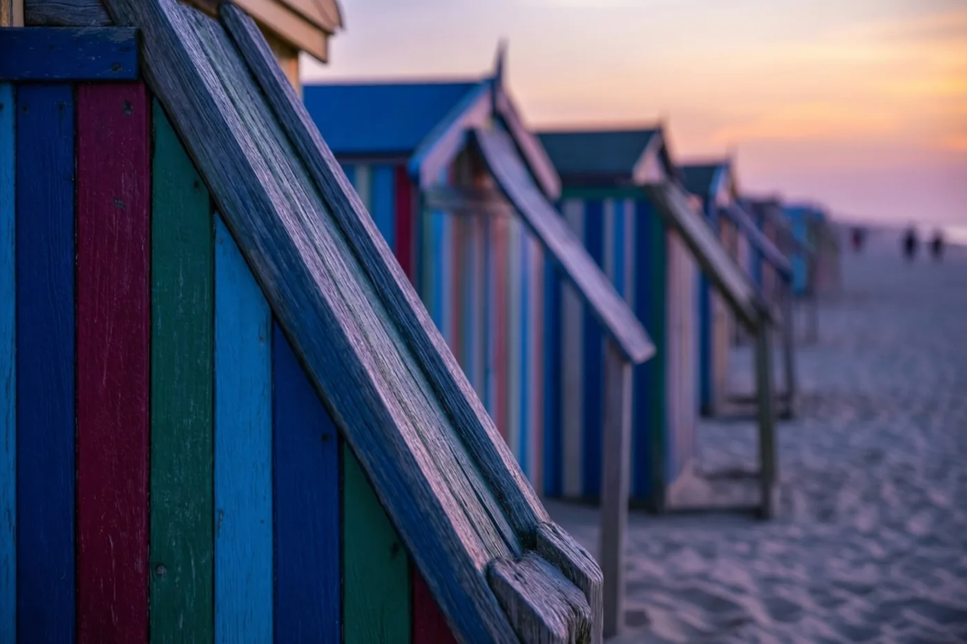 Kleurrijk gestreepte strandcabines staan op een zandstrand bij zonsondergang, onder een hemel met warme oranje en paarse tinten.