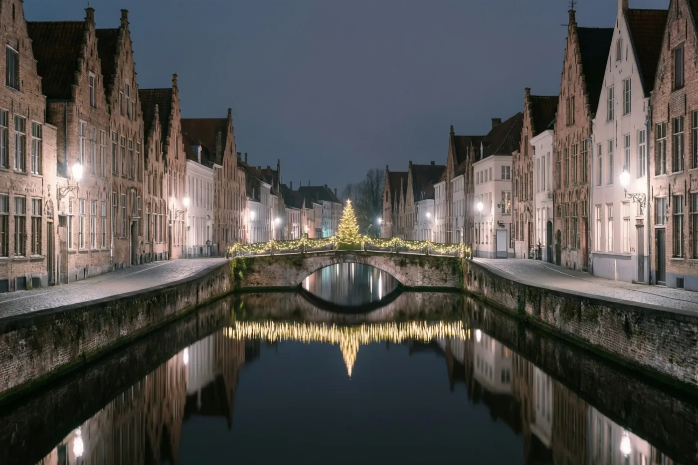 Historische gebouwen en een brug met kerstverlichting weerspiegelen in een waterloop onder de donkerblauwe nachthemel van een oud Vlaams stadje.
