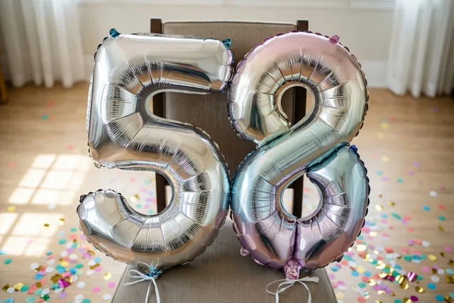 Glanzende cijferballonnen 58 met zilveren en wisselende pasteltinten hangen aan een stoel in een lichte kamer met confetti op de houten vloer.
