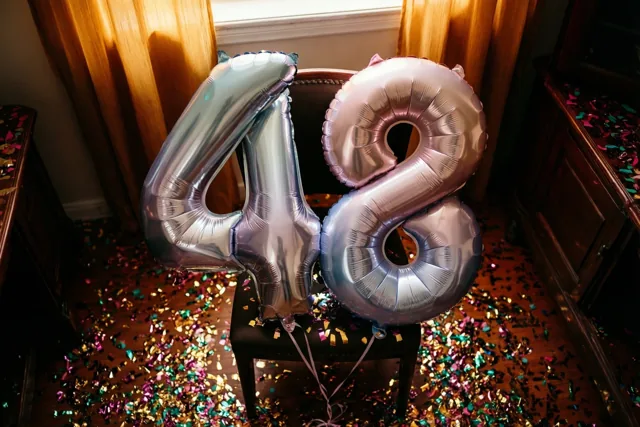 Twee grote glanzende ballonnen in de vorm van 4 en 8 zijn vastgebonden aan een stoel in een kamer met houten vloer, gordijnen en kleurrijke confetti in warm zonlicht.