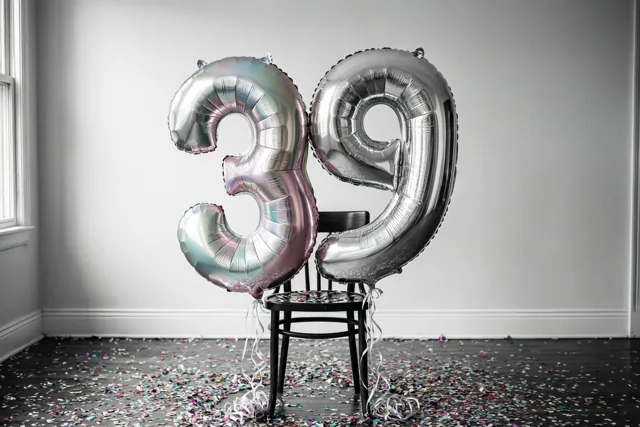 Twee grote cijferballonnen vormen 39 en zijn aan een zwarte stoel vastgemaakt in een lichte kamer, met een zilveren pastelglans en confetti op de donkere vloer.