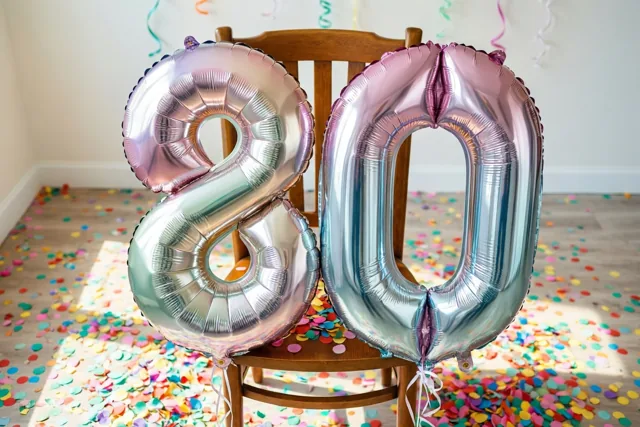 Twee grote metaalglanzende ballonnen vormen 80 aan een houten stoel in een lichte kamer met gekleurde confetti op de vloer en overwegend pastel- en zilvertinten.