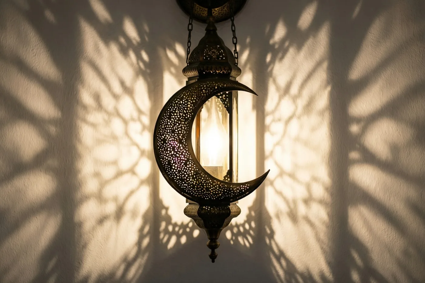 Een messing maansikkellantaarn met fijne uitsparingen hangt tegen een witte muur, met warm licht van binnenuit en een schaduw met patronen op de muur.