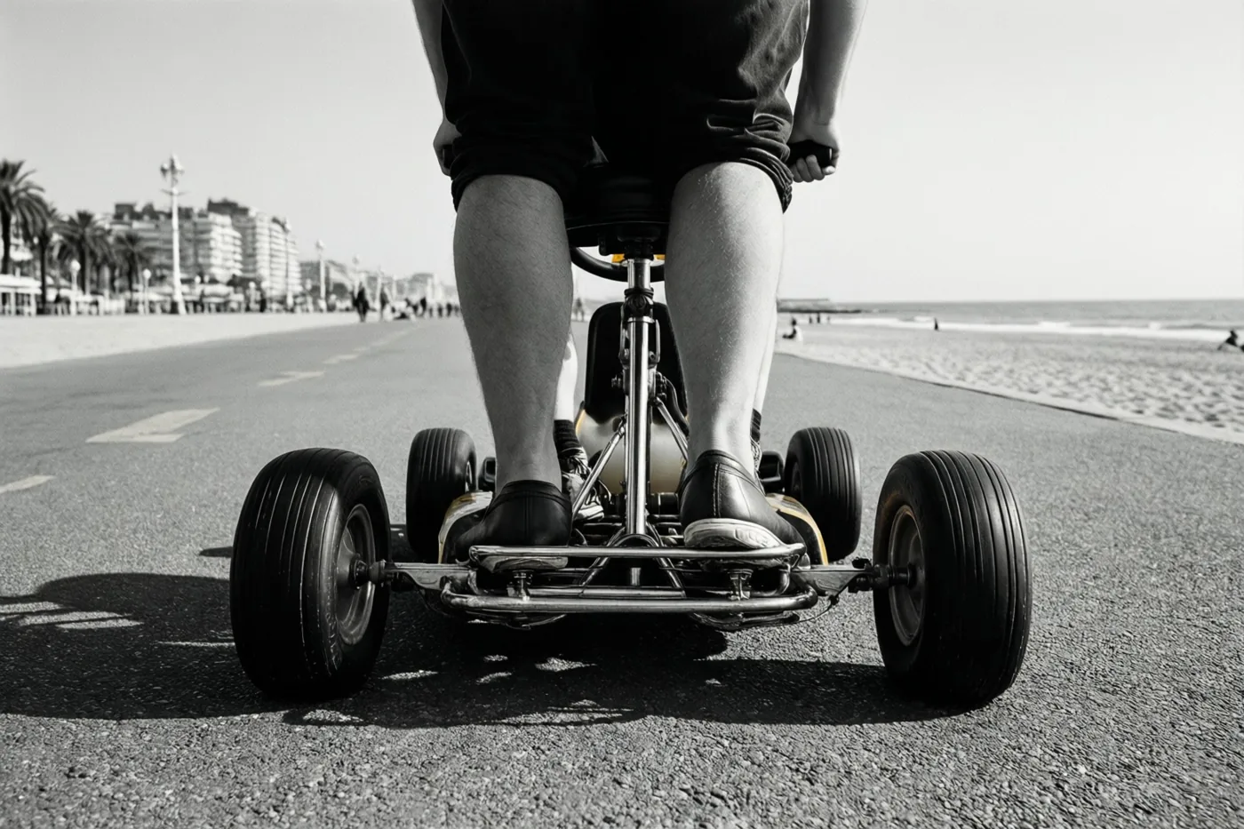 Een zwart-witfoto vanuit een laag standpunt toont de benen van iemand op een gocart op een verharde zeedijk, met rechts strand en zee en gebouwen in de verte.