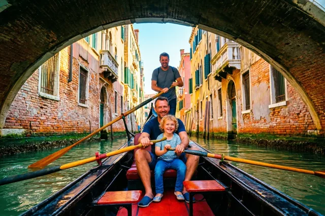 Een vader roeit een gondel met een rood interieur waarin een glimlachend kind zit, gezien onder een stenen brugboog met kleurrijke oude gevels langs het groene kanaalwater.