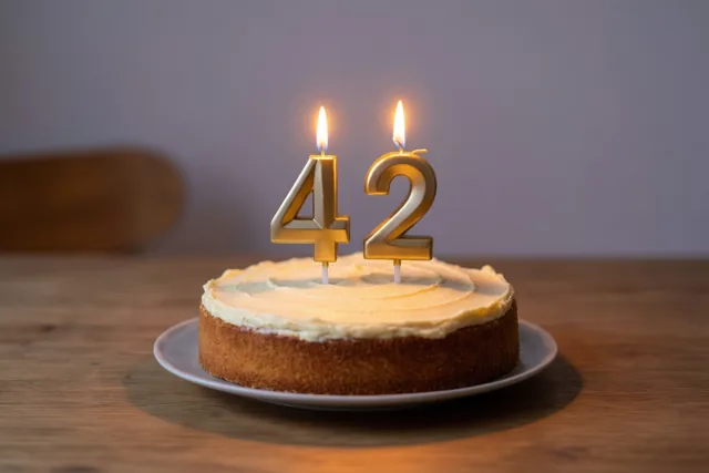 Een goudbruine zelfgemaakte roomtaart met brandende cijferkaarsjes die 42 vormen staat op een houten tafel in warm kaarslicht tegen een zachte achtergrond.