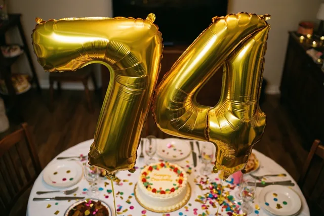 Gouden folieballonnen vormen 74 boven een ronde tafel met een versierde taart en kleurrijke confetti in een warm verlichte ruimte.