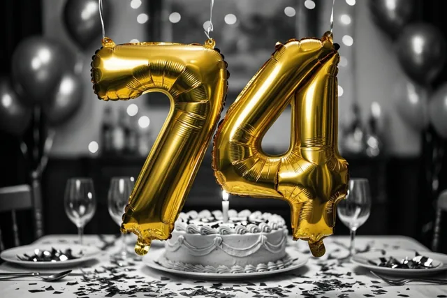 Gouden folieballonnen in de vorm van 7 en 4 hangen boven een tafel met taart, confetti en glazen tegen een zwart-witte achtergrond.