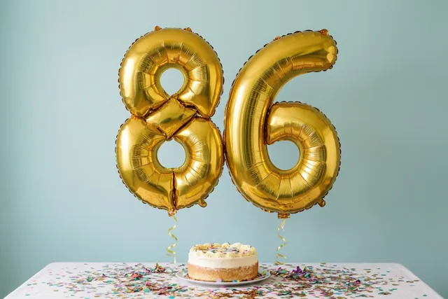 Gouden folieballonnen in de vorm van 8 en 6 zweven boven een tafel met een wit tafelkleed, een kleine taart en confetti tegen een effen lichtblauwe achtergrond.