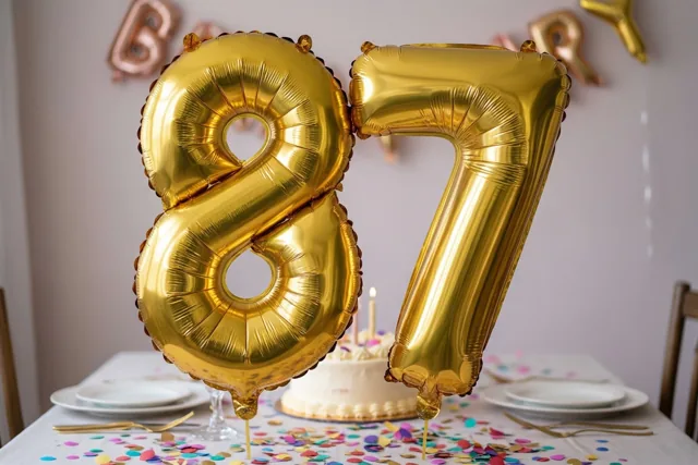 Grote glanzende gouden cijferballonnen vormen 87 boven een tafel met een taart, kleurrijke confetti en een licht tafelkleed.