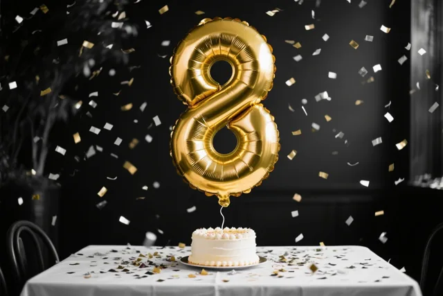 Een grote gouden ballon in de vorm van een 8 hangt boven een wit geglazuurde taart op een wit tafelkleed, met gouden en witte confetti tegen een donkere achtergrond.