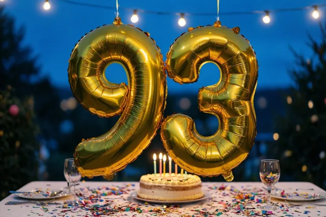 Gouden cijferballonnen vormen 93 boven een feesttafel buiten in de schemering, met lichtslingers, confetti en een taart met brandende kaarsjes.