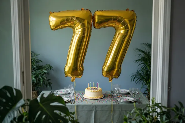 Twee gouden folieballonnen vormen 77 boven een feesttafel met taart, borden, glazen en confetti, met planten en een blauwgrijze muur op de achtergrond.
