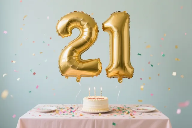 Gouden folieballonnen vormen het getal 21 boven een tafel met een roze kleed, confetti en een wit taartje met drie brandende kaarsen tegen een lichtblauwe achtergrond.