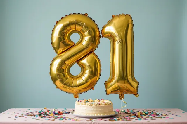 Grote gouden folieballonnen vormen 81 boven een tafel met een roze tafelkleed, taart en confetti, tegen een effen lichtblauwe muur.