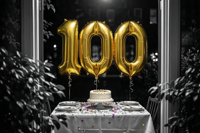 Drie grote gouden folieballonnen vormen 100 boven een tafel met taart en confetti, met planten rondom en een zwart-witte achtergrond bij nacht.