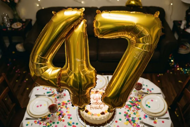 Grote glanzende gouden folieballonnen vormen 47 boven een feestelijk gedekte tafel met kleurrijke confetti en een verjaardagstaart in een warm verlichte kamer.