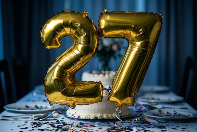 Gouden folieballonnen vormen 27 boven een tafel met gekleurde confetti, een witte taart en borden, voor een donkerblauw gordijn.