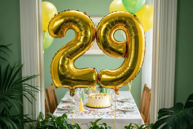 Gouden folieballonnen in de vorm van 29 hangen boven een tafel met taart, borden en confetti in een groene kamer met witte afwerking, gezien door een deuropening.