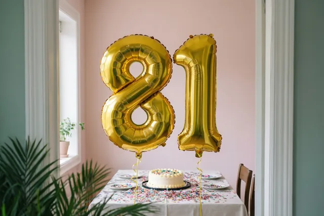 Twee grote gouden folieballonnen in de vorm van 8 en 1 zweven boven een tafel met taart en confetti in een kamer met roze muren en groene planten.