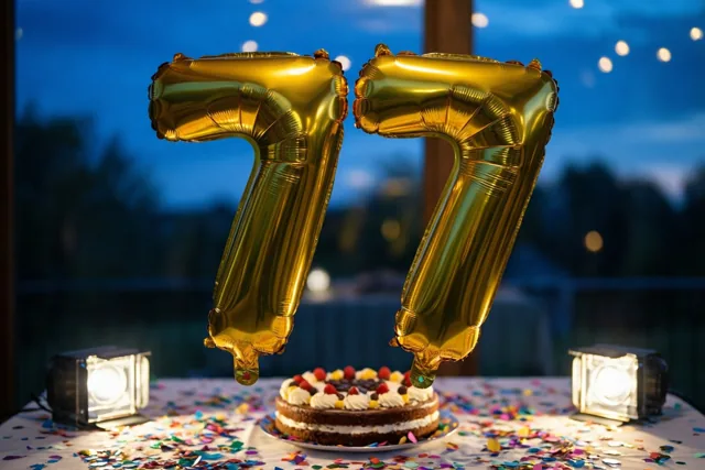 Gouden folieballonnen vormen 77 boven een tafel met kleurrijke confetti en een versierde taart, tegen een schemerlucht met lichtslingers.