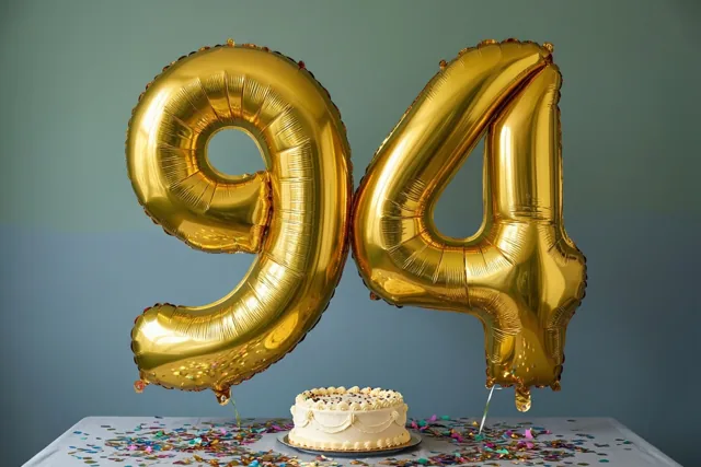Gouden folieballonnen vormen 94 boven een tafel met kleurrijke confetti en een kleine witte taart tegen een blauwgroene achtergrond.