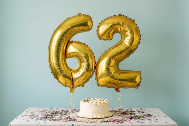 Gouden folieballonnen in de vorm van 6 en 2 zweven boven een witte tafel met confetti en een kleine witte taart tegen een lichtblauwe achtergrond.