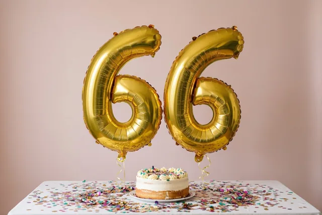 Twee grote glanzende gouden folieballonnen vormen 66 boven een witte tafel met gekleurde confetti en een kleine taart tegen een effen roze achtergrond.