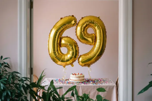 Grote glanzende gouden folieballonnen vormen 69 boven een tafel met confetti en een kleine taart, tegen een lichtroze muur met groene planten op de voorgrond.