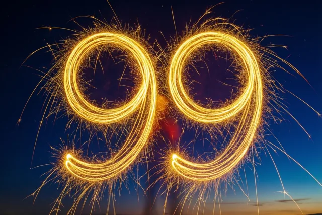 Gouden lichtsporen van sterretjes vormen 99 tegen een donkerblauwe nachtelijke hemel met oranje schemerlicht aan de horizon.