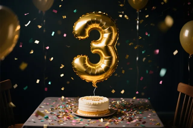Een grote gouden cijferballon 3 zweeft boven een taart op een tafel vol confetti, tegen een donkere achtergrond met zwevende confetti en gouden ballonnen.