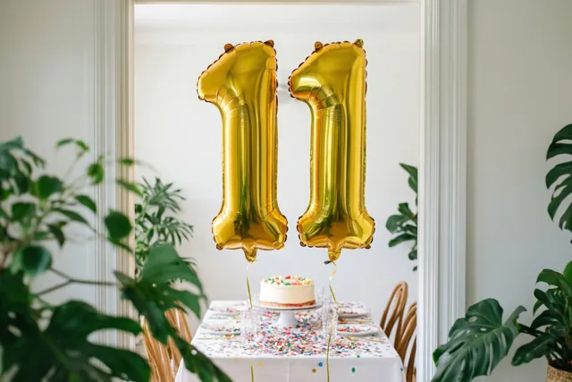 Gouden folieballonnen vormen 11 boven een tafel met confetti en taart, omlijst door een deuropening met witte randen en groene planten aan weerszijden.