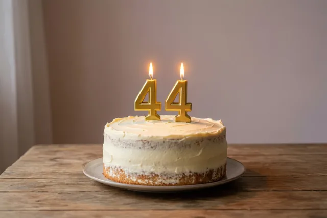 Een zelfgemaakte roomtaart met twee brandende gouden cijferkaarsjes die 44 vormen staat op een houten tafel in warm kaarslicht.