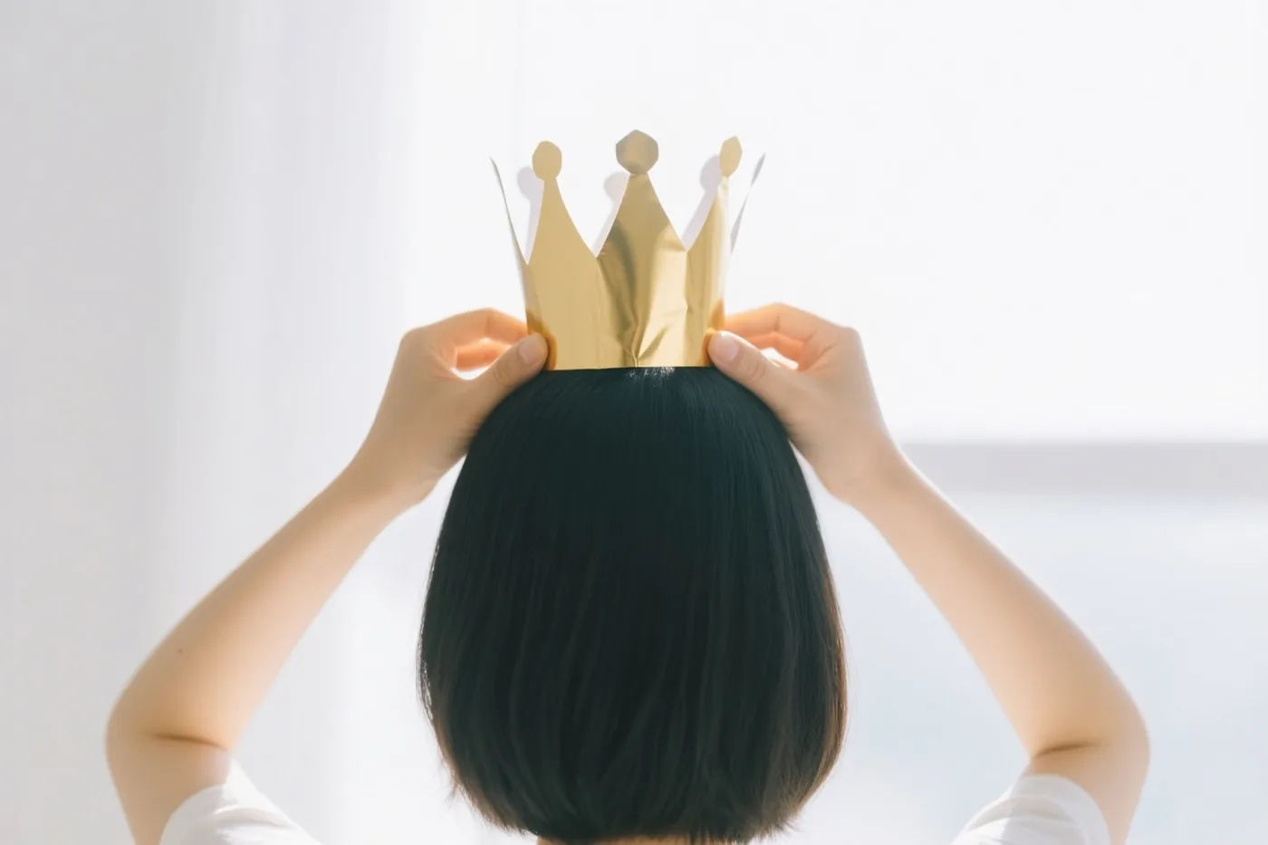 Twee handen plaatsen een goudkleurige papieren kroon op het hoofd van een persoon met kort zwart haar, van achteren gezien tegen een helderwitte achtergrond.