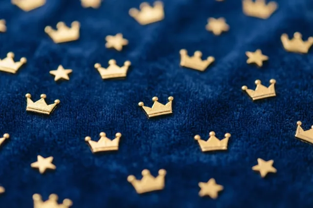 Kleine gouden kroontjes en sterren liggen verspreid op diepblauw fluweel, met een sterk contrast tussen het goud en het blauw.