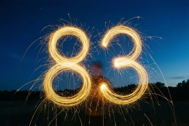 Gouden lichtsporen van sterretjes vormen 83 tegen een donkerblauwe nachthemel, met silhouetten van bomen op de achtergrond.