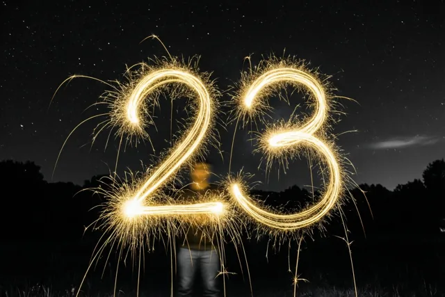 Gouden lichtsporen vormen 23 tegen een sterrenhemel, met donkere silhouetten van bomen op de achtergrond.