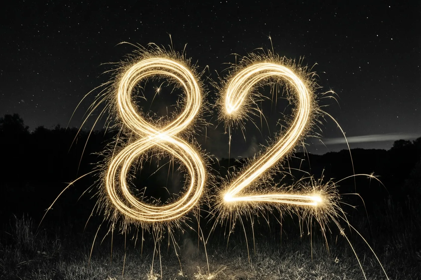 Gouden lichtsporen van sterretjes vormen het getal 82 boven gras, met een sterrenhemel en donkere boomsilhouetten op de achtergrond.